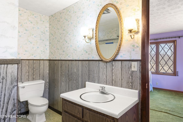 half bathroom featuring a wainscoted wall, toilet, vanity, a textured ceiling, and wallpapered walls