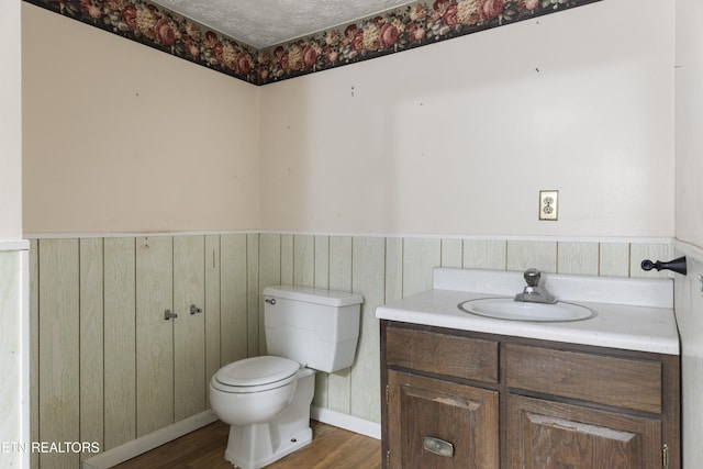 half bath with toilet, wainscoting, a textured ceiling, vanity, and wood finished floors