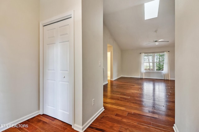 unfurnished room with ceiling fan, vaulted ceiling with skylight, dark wood-type flooring, and baseboards