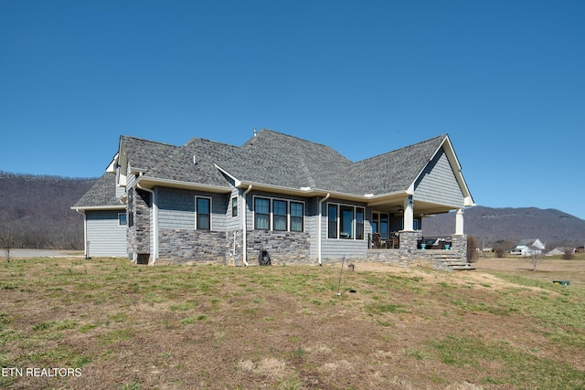 back of house with a shingled roof, stone siding, a mountain view, and a lawn