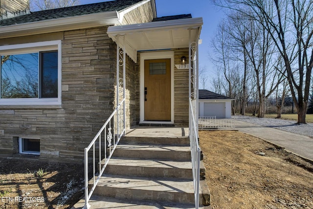 entrance to property featuring stone siding, driveway, a detached garage, and roof with shingles