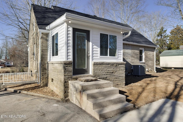 view of exterior entry featuring stone siding, cooling unit, and a shingled roof