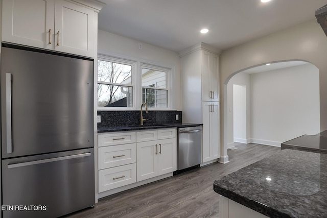 kitchen with dark stone counters, appliances with stainless steel finishes, arched walkways, white cabinetry, and a sink