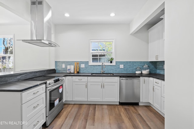 kitchen featuring island exhaust hood, wood finished floors, dark countertops, appliances with stainless steel finishes, and a sink