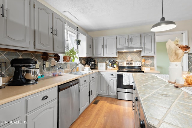 kitchen featuring light wood finished floors, tile counters, stainless steel appliances, under cabinet range hood, and a sink