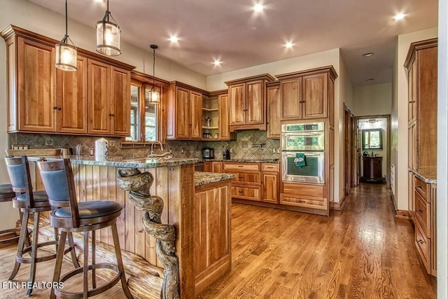 kitchen featuring double oven, a peninsula, a kitchen breakfast bar, brown cabinets, and dark stone countertops