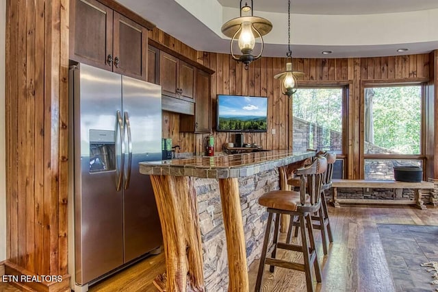 kitchen featuring a breakfast bar area, wooden walls, stainless steel refrigerator with ice dispenser, dark countertops, and dark wood finished floors