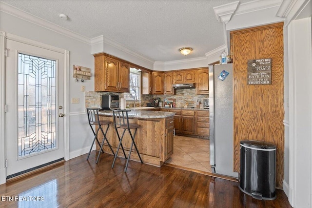 kitchen with tasteful backsplash, brown cabinets, freestanding refrigerator, a peninsula, and under cabinet range hood