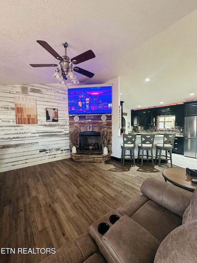 living area with a textured ceiling, a stone fireplace, wood finished floors, and a ceiling fan