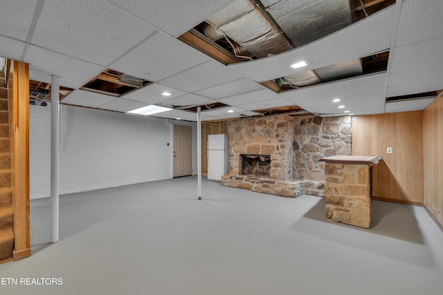 finished basement featuring a stone fireplace, wood walls, a drop ceiling, and freestanding refrigerator