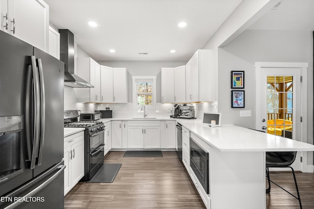 kitchen with a breakfast bar area, a peninsula, a sink, appliances with stainless steel finishes, and wall chimney range hood