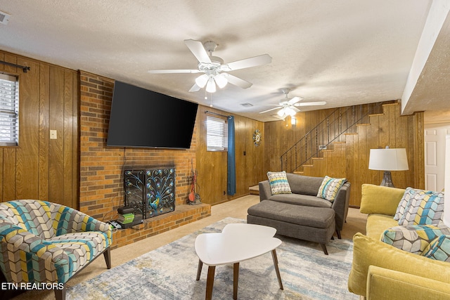 carpeted living room featuring a brick fireplace, ceiling fan, wooden walls, a textured ceiling, and stairs