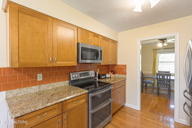 kitchen with brown cabinetry, light wood-style flooring, appliances with stainless steel finishes, light stone counters, and backsplash