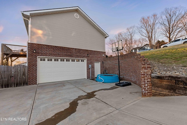 property exterior at dusk featuring a garage, brick siding, fence, concrete driveway, and a hot tub