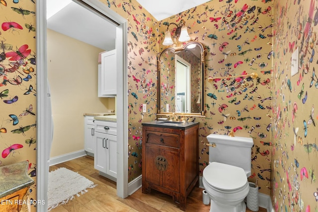 bathroom featuring baseboards, toilet, vanity, and wallpapered walls