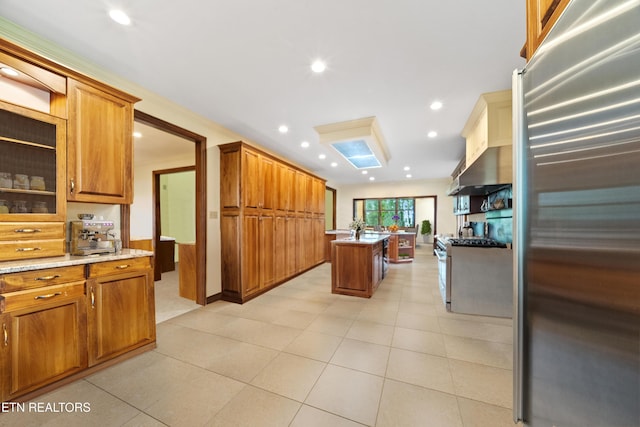 kitchen featuring stainless steel refrigerator, brown cabinetry, recessed lighting, under cabinet range hood, and a center island with sink