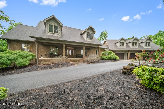 new england style home featuring a garage, covered porch, brick siding, a shingled roof, and driveway