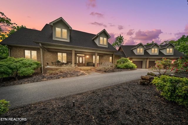 cape cod home with a porch, a garage, driveway, and brick siding