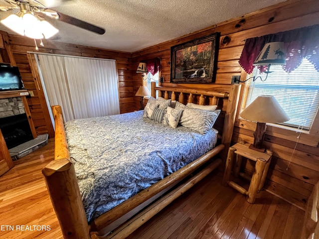 bedroom featuring a ceiling fan, a stone fireplace, wooden walls, a textured ceiling, and hardwood / wood-style floors