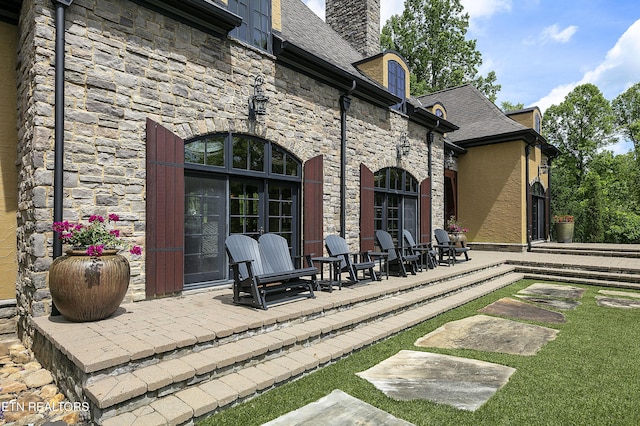 rear view of property with stone siding, roof with shingles, stucco siding, a chimney, and a patio area