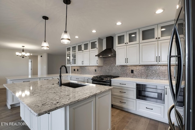 kitchen featuring black appliances, an island with sink, a sink, wood finished floors, and wall chimney exhaust hood