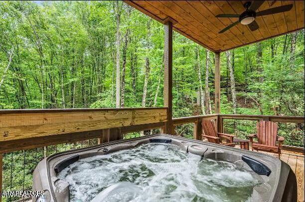 wooden terrace featuring an outdoor hot tub, a ceiling fan, and a view of trees