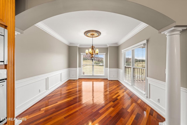 unfurnished dining area featuring arched walkways, decorative columns, an inviting chandelier, wainscoting, and hardwood / wood-style floors