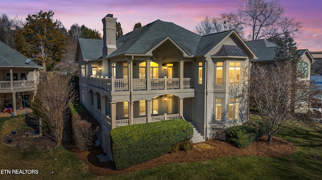 rear view of property featuring a shingled roof, a chimney, a balcony, and stucco siding