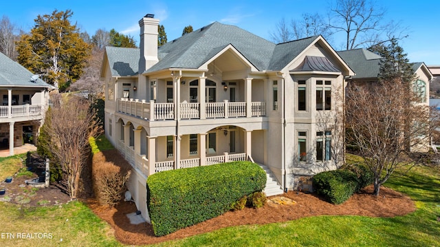 back of house with a shingled roof, a chimney, a balcony, and stucco siding