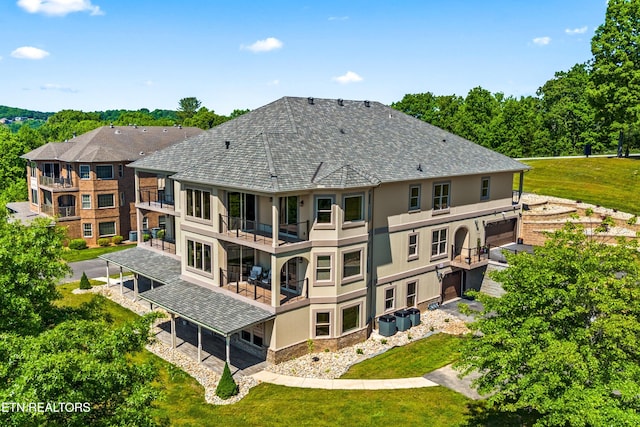 rear view of house featuring a shingled roof, a balcony, central AC unit, and stucco siding