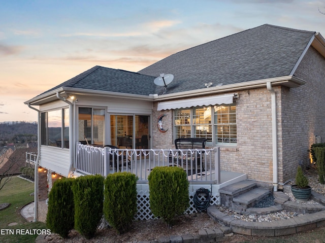 back of house at dusk featuring brick siding and roof with shingles