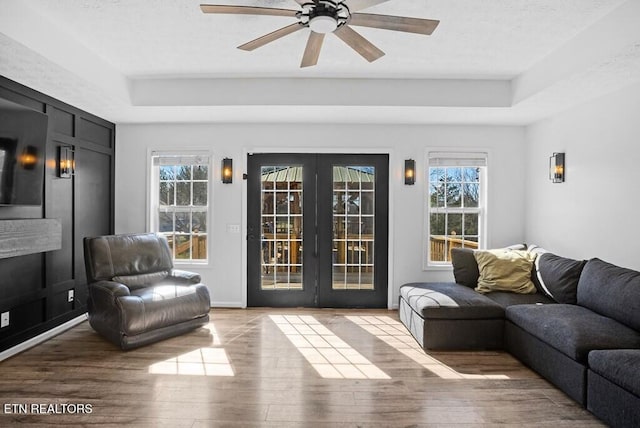 living area with ceiling fan, a tray ceiling, wood finished floors, and french doors