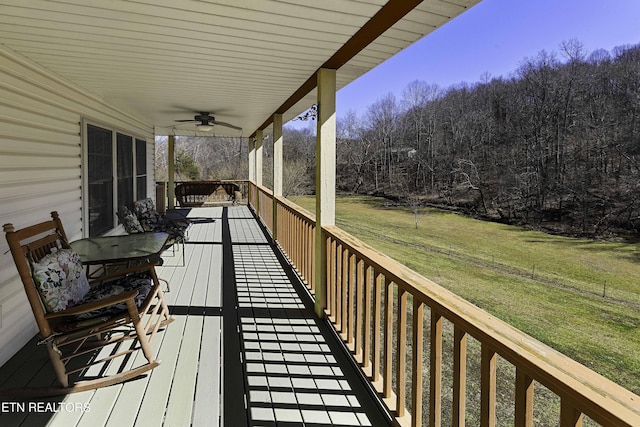 wooden deck with a porch, ceiling fan, a forest view, and a lawn