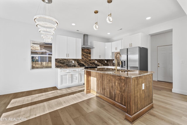 kitchen with stainless steel refrigerator with ice dispenser, light wood finished floors, decorative backsplash, dark stone countertops, and wall chimney exhaust hood