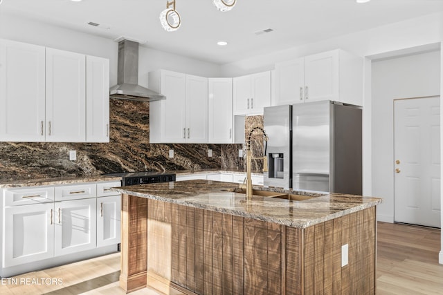 kitchen with a sink, wall chimney range hood, stainless steel fridge with ice dispenser, tasteful backsplash, and dark stone countertops