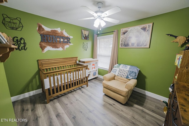 bedroom with a ceiling fan, a nursery area, baseboards, and wood finished floors