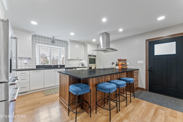 kitchen with light wood finished floors, dark countertops, a kitchen breakfast bar, island exhaust hood, and stainless steel appliances