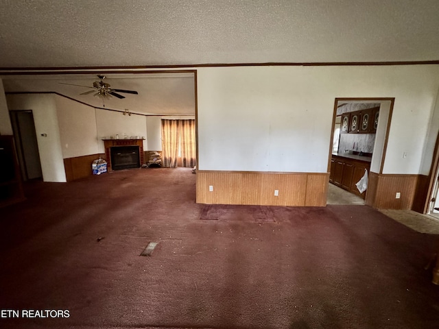 unfurnished living room featuring a wainscoted wall, a fireplace, ceiling fan, wooden walls, and a textured ceiling