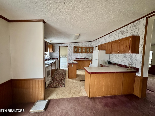 kitchen featuring brown cabinetry, wainscoting, washer / dryer, white appliances, and a peninsula