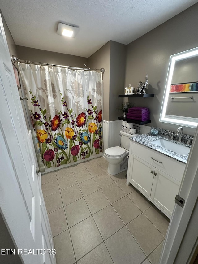 bathroom featuring a textured ceiling, toilet, vanity, wainscoting, and tile patterned floors