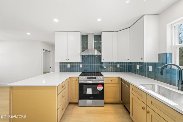 kitchen featuring appliances with stainless steel finishes, a sink, light wood-type flooring, a peninsula, and wall chimney exhaust hood