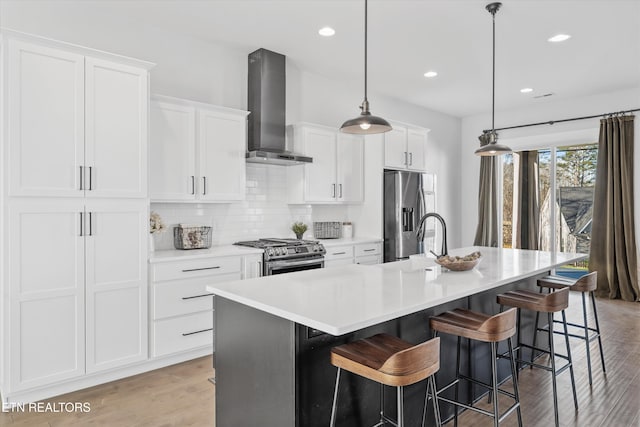 kitchen featuring an island with sink, a sink, stainless steel appliances, white cabinets, and wall chimney exhaust hood