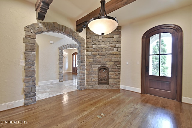 entrance foyer featuring arched walkways, beamed ceiling, a stone fireplace, and wood finished floors