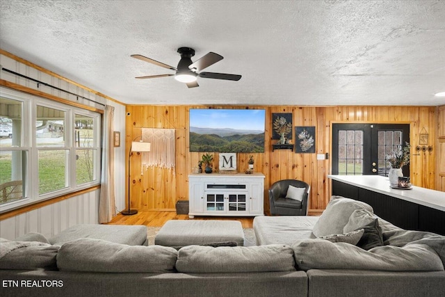 living area with a textured ceiling, wooden walls, wood finished floors, a ceiling fan, and french doors