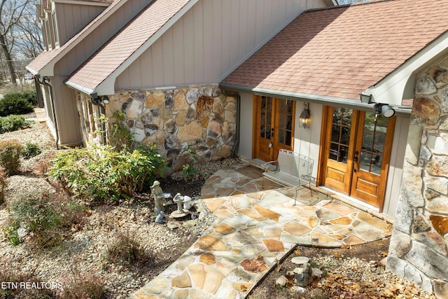 view of exterior entry featuring french doors, stone siding, and roof with shingles