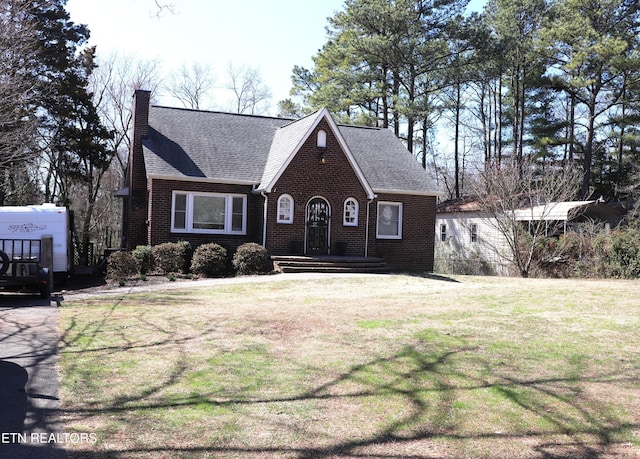 english style home with brick siding, a chimney, a front lawn, and roof with shingles