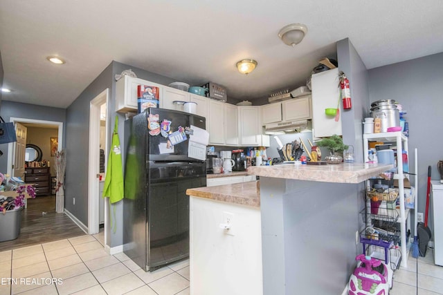 kitchen with a peninsula, under cabinet range hood, light tile patterned flooring, and freestanding refrigerator