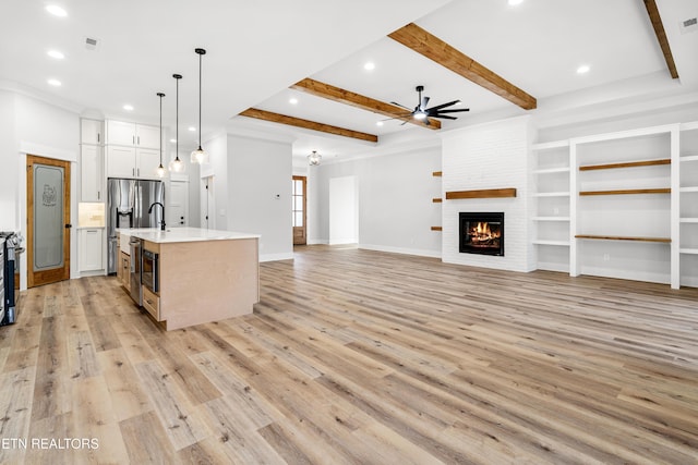 kitchen with light wood finished floors, light countertops, a brick fireplace, a kitchen island with sink, and beamed ceiling