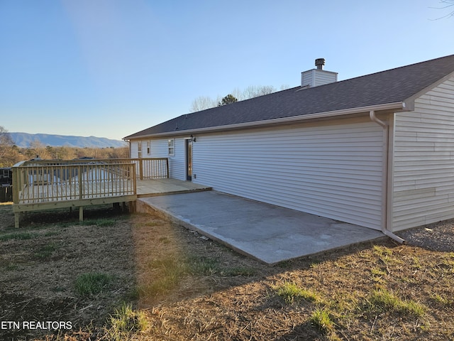 back of house featuring a deck with mountain view, a patio area, roof with shingles, and a chimney