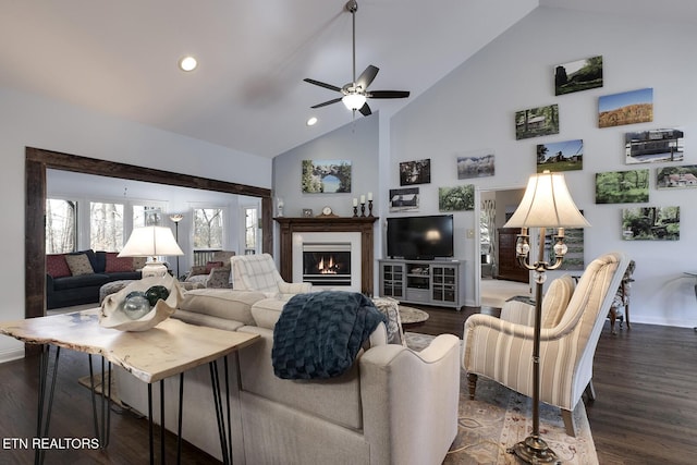 living area with a warm lit fireplace, high vaulted ceiling, ceiling fan, baseboards, and dark wood-style floors